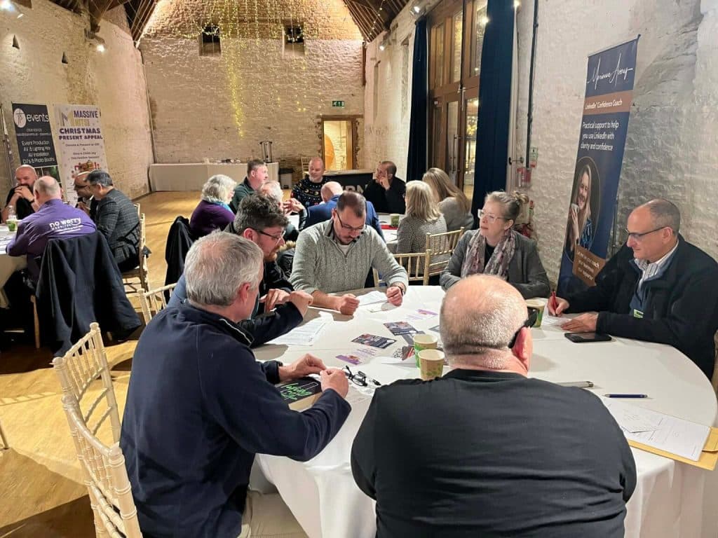 People sit around round tables in a rustic, high-ceilinged room with white stone walls, engaged in discussion and writing on papers. Banners and informational material suggest an active Business Networking event is under way.