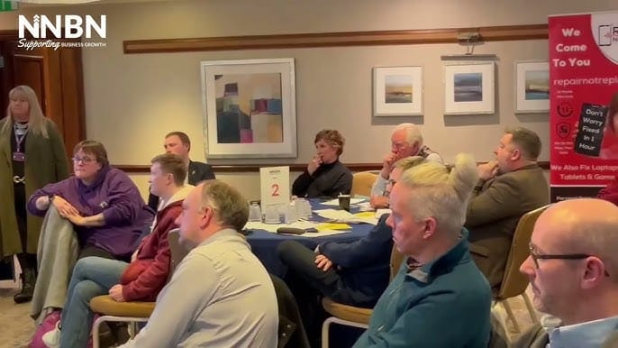 A group of people sit attentively around tables in a conference room, listening to a speaker. Some have notepads, and business networking banners are visible on the walls. The NNBN logo appears in the top left corner.