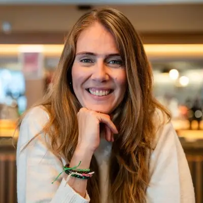 A woman with long light brown hair smiles at the camera, resting her chin on her hand. She wears a light jumper and colourful bracelets, seated indoors with a softly blurred background.