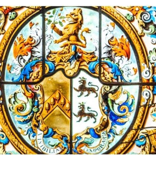 Colourful stained glass window featuring a crest with a lion on top, two lions inside a shield, swirling floral patterns, and the Latin motto Visibilia Temporaria.