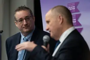 Two men in suits speak at an event. One man holds a microphone whilst gesturing, and the other listens attentively. Award banners are visible in the blurred background.