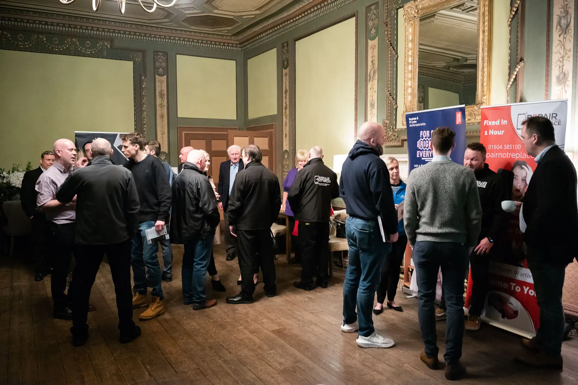 A group of people, mostly men, stand and talk in a large ornate room with wooden floors. Some face stands with banners and posters, suggesting a networking or recruitment event. Chandeliers and gold-framed mirrors decorate the space.